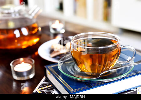 Teatime Szene mit Glas Teekanne und Tasse Tee, altes Buch und heißen Dampf. Idyllische Tea Time oder Entspannung Szene mit Plätzchen und Kerzen. Stockfoto