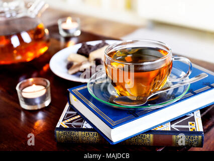 Teatime Szene mit Glas Teekanne und Tasse Tee, altes Buch und heißen Dampf. Idyllische Tea Time oder Entspannung Szene mit Plätzchen und Kerzen. Stockfoto