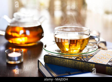 Teatime Szene mit Glas Teekanne und Tasse Tee, altes Buch und heißen Dampf. Idyllische Tea Time oder Entspannung Szene mit Plätzchen und Kerzen. Stockfoto