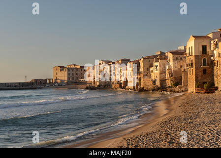 Alte Häuser am Meer auf den Klippen der Küste von Sizilien gebaut und neben einem leeren Strand, Baden in der letzten, warmen Abendlicht in Cefalù in Italien. Stockfoto