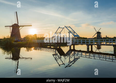 Eine Landschaft in den Niederlanden mit zwei Windmühlen und eine Zugbrücke in die niedrige Sonne bei Sonnenuntergang mit Reflexion im Wasser in Kinderdijk, Niederlande. Stockfoto