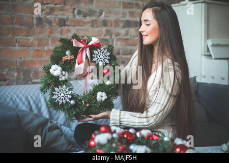 Glückliche junge floristin Frau am häuslichen Arbeitsplatz sitzen und Weihnachten immergrüner Baum Kranz mit roten Kugeln aus Glas, Kegel, Farbband und Zimt. Real Stockfoto