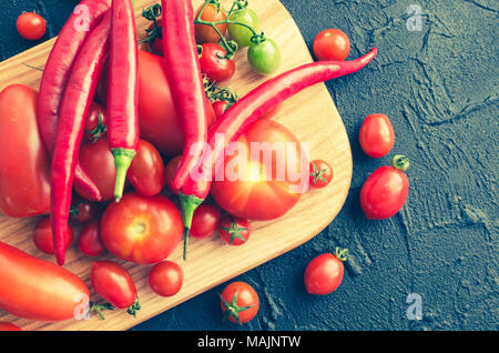 Verschiedenen bunten Tomaten und red hot chili peppers auf Holzbrett auf dunklen Tisch aus Stein. Verschiedene Arten von frischen Bio Tomaten auf schwarzem Beton bac Stockfoto
