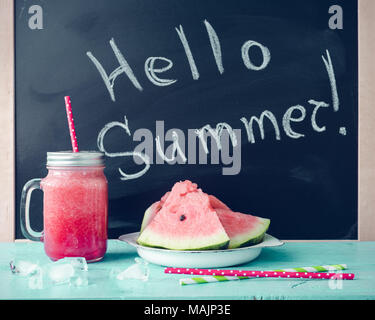 Hallo Sommer auf der Tafel mit Wassermelone Saft im Glas Marmeladenglas mit Scheiben von Wassermelone und Eis auf blauem Hintergrund geschrieben. Frisches Obst flav Stockfoto