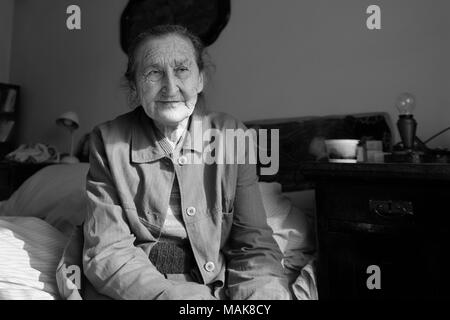 Schöne 80plus-jährigen Senior Frau Porträt. Schwarz-weiß Bild von älteren Menschen sorgen Frau auf einem Bett in einem Pflegeheim sitzen. Stockfoto