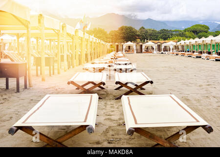 Tageslicht, beige, Liegestühlen und Sonnenschirmen am Strand. Umkleideräume, große Berge und bewölkter Himmel im Hintergrund. Negative Kopie Raum, Ort für Te Stockfoto