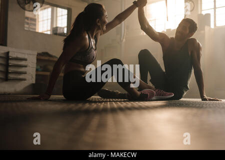 Fitter Mann und Frau sitzen auf dem Boden und High Fiving im Fitnessstudio. Fitnesspaar nach erfolgreichem Workout im Fitnessstudio. Stockfoto