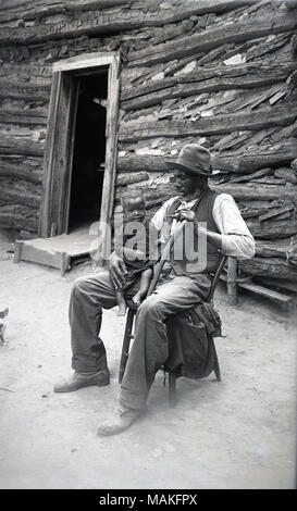 Vertikale, schwarze und weiße Blick auf einen älteren Mann, stützte sich auf seinen Stock, sitzt vor einer Hütte mit einem kleinen Kind auf seinem Schoß. Es ist eine offene Tür Weg hinter sich. Das Kind erscheint Rachitis oder ein Bein Deformität zu haben. Titel: Robert Goodin, Vigus, Missouri, St. Louis County. . Um 1910. Oscar Kuehn Stockfoto