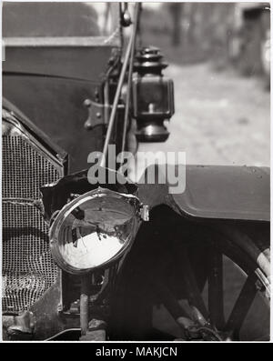 Vertikal, schwarz-weiß Foto zeigt eine Nahaufnahme eines beschädigten Auto Scheinwerfer und Grill. Scheinwerfer, rechts Der Wagen wurde aus Platz heraus geklopft worden, und der Grill hat eine Delle in der rechten unteren Ecke. Der Fotograf mit seiner Kamera und Stativ gesehen werden kann im Scheinwerfer wider. Für das vollständige Bild, siehe P 0054-00025. Titel: Nahaufnahme eines beschädigten Auto Scheinwerfer. . Um 1910. Holt, Charles Clement, 1866-1925 Stockfoto