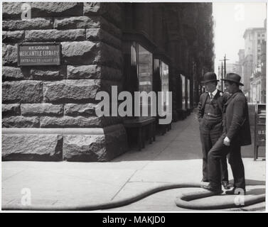 Horizontal, schwarz-weiß Foto zeigt die Ecke der St. Louis Mercantile Gebäude der Bibliothek. Ein Schild auf dem Gebäude lautet: t. Louis Mercantile Bibliothek/Eingang auf Locust St.' mehrere Vitrinen können entlang der Seite des Gebäudes gesehen werden, die sich in den Hintergrund. Zwei Männer stehen auf der Straße Ecke Tabakpfeifen. Einer der Männer seinen Fuß auf einen großen Schlauch dem schlangenpfad Bürgersteig am unteren Bildrand. Titel: Ecke der St. Louis Mercantile Bibliothek Gebäude, 510 Locust Street. . Zwischen 1900 und 1920. Holt, Charles Clement, 1866-1925 Stockfoto