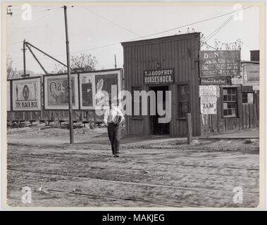 Horizontal, schwarz-weiß Foto zeigt einen Jungen oder jungen Mann zu Fuß entfernt von T.J. Godfrey Horseshoer, 3968 Chouteau entfernt. Der Junge ist, was zu sein scheint eine der Zeitungen, die Stapel, und er ist über zwei Sätze von Straßenbahn Spuren, die durch den Vordergrund laufen zu überqueren. T.J. Godrey's ist ein einstöckiges Gebäude aus Holz, mit Werbung zugepflastert entlang der Seite. Mehrere große Plakate werden auf der rechten Seite des Gebäudes. Werbung fördern die Black Box, Oasis Zigaretten, Jag. Alders sanitär Dienstleistungen, Pillsbury ist am Besten, Mehl und King's Theatre. Titel: T.J. Godfrey Horseshoer Stockfoto