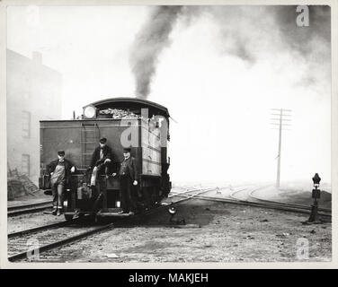 Horizontal, schwarz-weiß Foto zeigt drei Männer auf der Rückseite einer steam-engine in einem Rail Yard. Zwei der Männer stehen auf einem Trittbrett, während die dritte auf einer Schiene oder Rohr thront. Ein Mann kann die Leiter sein, da er trägt eine einheitliche Hut und Anzug, während die beiden anderen Herren Bauarbeiter Schutzkleidung tragen. Ein Scheinwerfer ist an der Spitze einer Kurze Leiter und Kohle angehäuft im Bunker kann Dahinter gesehen werden. Eine Wolke aus Rauch aufsteigt, vom Bahnhof. Mehrere Eisenbahnschienen kreuz und quer im Hintergrund. Eine handschriftliche Notiz auf der Rückseite des Drucks, wahrscheinlich von der Schule Schwestern von Notre D Stockfoto