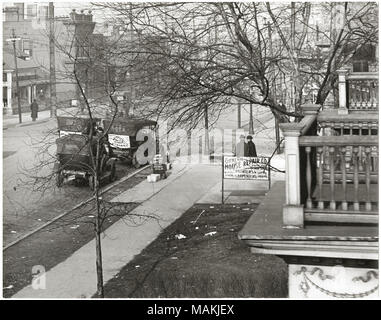 Horizontal, schwarz-weiß Foto zeigt ein nicht identifizierter Bürgersteig Szene. Der Gehweg verläuft diagonal durch die Mitte des Bildes, und zwei Männer kann stehend in der Nähe der Straße Ecke gesehen werden. Zwei 1910-Ära Autos sind entlang der Bordstein geparkt, einer mit einem Schild mit Werbung einen Optiker. Zwei Balkone Projekt in der rechten Seite des Fotos, und ein Schild vor dieser Gebäude wirbt für die Allgemeine Haus reparieren Unternehmen. Mehrere Gebäude auf der anderen Straßenseite sind sichtbar auf der linken Seite des Bildes. Titel: Nicht identifizierte Bürgersteig Szene mit Balkonen, geparkte Autos und Fußgänger. . B Stockfoto