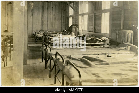 Horizontale, sepia Foto, das Innere eines Holz-seitig Gebäude, wahrscheinlich als Gehäuse für Soldaten dienen. Zwei Reihen von Kinderbetten Linie die Außenwände der Gebäude. Die meisten Betten sind gemacht, und viele der Babybetten mess Kits oder Tassen auf Sie. Schreiben Sie auf der Rückseite des Fotos gefunden werden, aber die Schrift ist teilweise verdeckt von scrapbook Papierreste. Titel: Anzeigen der Zeilen von Kinderbetten in einem Wood-Sided Kaserne Gebäude. . Zwischen ca. 1914 und ca. 1918. Michel, Carl Stockfoto