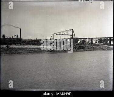 Horizontal, Schwarz und Weiß Foto, nicht identifizierte Industrieanlagen. Die Industrieanlagen gehören gestapelt Stapel von Protokollen, eine Hochbahn, und in niedrigen Gebäuden mit Schornsteinen im Hintergrund. Das Foto wurde aus über einen Fluss, der der Vordergrund füllt. Titel: Nicht identifizierte Industrieanlagen. . Zwischen 1900 und 1920. Stockfoto