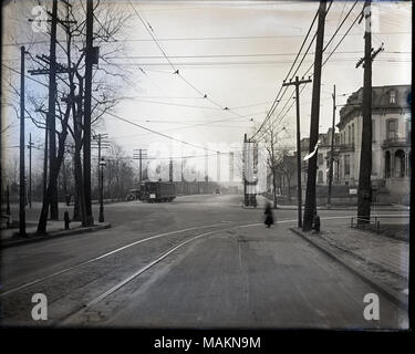 Horizontal, schwarz-weiß Foto zeigt eine Straße mit einer Straßenbahn, die möglicherweise an der Ecke der Mississippi und Lafayette. Das Foto wurde in einen 4-fach-Kreuzung. Eine der Straßenbeleuchtung hat ein Schild mit der Aufschrift 'Mississippi', aber die andere Straße nicht identifiziert. Es gibt Straße Auto Spuren durch den Vordergrund laufen, und eine verschwommene Frau über die Straße. Auf der linken Seite eine Straße Auto fährt ein Auto vorbei und viele Bäume hinter einem schmiedeeisernen Zaun, möglicherweise Lafayette Park. Auf der rechten Seite, Häuser säumen die Straße. Titel: Street Scene mit einer Straßenbahn, möglicherweise an der Stockfoto