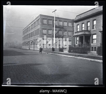 Horizontal, Schwarz und Weiß Foto, ein 3-stöckiges Werk oder Lager auf der gegenüberliegenden Straßenseite aus einem 2-stöckigen Mehrfamilienhaus Wohnhaus. Die Fabrik verfügt über große Fenster und ein Feuer - Flucht an der Seite des Gebäudes. Die Residence ist ein 2-stöckiges Ziegelgebäude, wahrscheinlich ein Mehrfamilienhaus und verfügt über eine kleine Veranda. Ein kleines Plakat mit einem hellen Hintergrund und ein Kreuz in der Mitte ist in einem der Fenster angezeigt wird, ist möglicherweise ein rotes Kreuz Service Flagge, während des Ersten Weltkrieges eine Straße mit Kopfsteinpflaster läuft durch den Vordergrund und zwei fussgänger gesehen werden kann. Original Umschlag wurde La Stockfoto