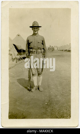 Vertikale, sepia Foto, ein Mann, der eine Uniform mit einem Gewehr für ein Bild in einem großen offenen Bereich posieren. Zelte können im Hintergrund des Bildes zu sehen. Titel: Uniformed Soldat posieren für ein Foto in ein Militärlager. . Zwischen ca. 1916 und ca. 1918. Stockfoto