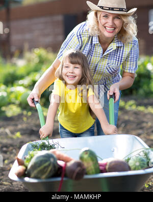 Adorable kind Mädchen und ihre Mutter schieben Schubkarre mit Gemüse an einem sonnigen Tag. Sommer arbeitet im Garten. Kind Mama hilft. Stockfoto