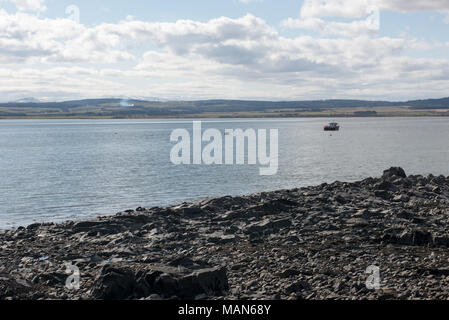 Felsige Küste mit ruhigem Meer und weit entferntem Boot auf Holy Island, Northumberland, Großbritannien, unter einem hellen Himmel mit Wolken und sanftem Licht. Stockfoto