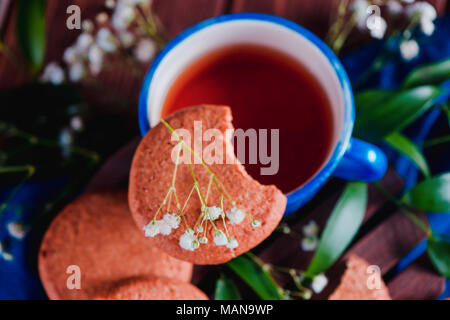 Nahaufnahme einer Teetasse mit Haferflocken Cookies und Feder gypsophila Blumen auf einem warmen Holz- Hintergrund. Blau Keramiktasse auf blauem Leinen Serviette. Breakfas Stockfoto