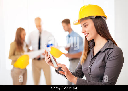 Lächelnden jungen Frau Architekt mit Helm im Büro und mit digitalen Tablet. Ihre Kollegen sprechen im Hintergrund. Stockfoto