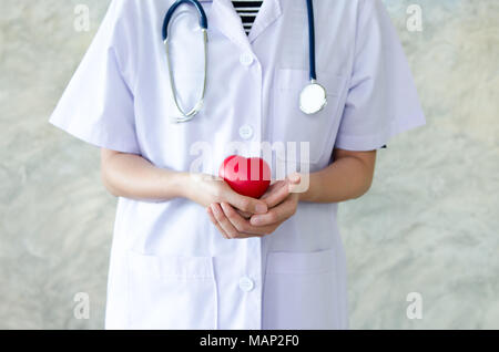 Close up junge Frau Doktor zwei Hände halten roten Spielzeug Herz. Hände halten ein rotes Herz als Symbol für Liebe und Sorgfalt. Gesundheitswesen und medizinische Konzept Stockfoto