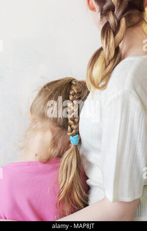 Ein junges Mädchen mit einem Zopf Umarmungen ein kleines Mädchen mit einem langen Licht Braid, stehen Sie mit dem Rücken zu der Kamera ältere Schwester Pflege und Liebe. Stockfoto