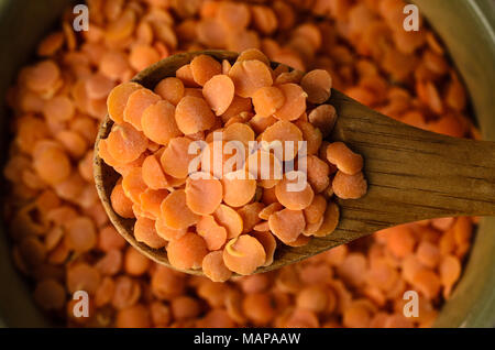 Overhead close up (Makro) von einer hölzernen Löffel gefüllt mit roten Linsen und in Richtung Betrachter aufgehoben. Volle Schüssel in Soft Focus Hintergrund unten. Stockfoto