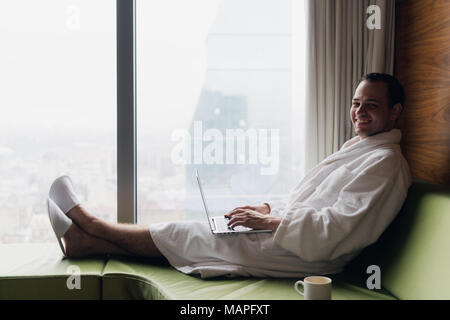 Junge lächelnde Geschäftsmanns, der auf dem Laptop Computer tragen weiße Bademantel saß in der Nähe der Fenster mit Tasse Kaffee bei Sonnenaufgang Blick auf die Stadt. Motivation Konzept Stockfoto