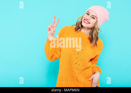 Crazy wunderschöne trendige Mädchen in Senf farbigen Pullover und rosa Mütze Hut Making Peace Sign Geste. Attraktive cool junge lächelnde Frau. Stockfoto