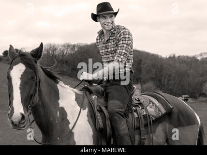 Schöner Mann, männlich Cowboy im Sattel sitzen auf Pferd mit Hügel und Bäume im Hintergrund Stockfoto