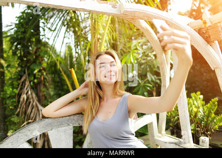 Junge Frau nutzt Smart Phone für selfie an einem sonnigen Tag, Hintergrund Sonnenschein grüne Palmen in Thailand, Phuket. Konzept der neuen Technologien für eine bessere l Stockfoto