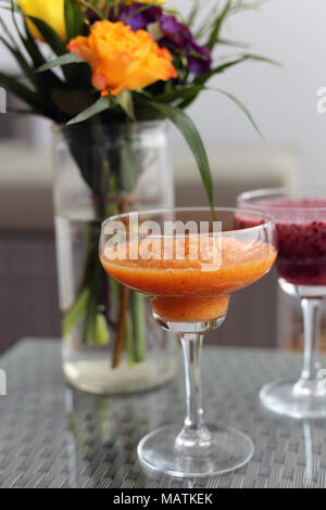 Zwei selbstgemachte DIY Smoothies (orange und lila Berry Smoothie) auf einem Balkon Tisch mit bunten Blumen im Hintergrund. Gesund und lecker! Stockfoto