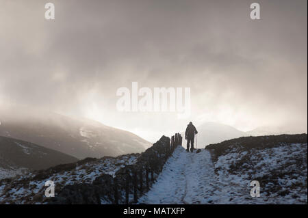 Ein Mann Wandern im Schnee mit der Hilfe von einem Stock auf einem Pfad in den Winter Under A Dark Sky Stockfoto