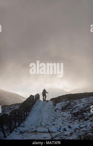 Ein Mann Wandern im Schnee mit der Hilfe von einem Stock auf einem Pfad in den Winter Under A Dark Sky Stockfoto