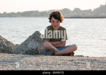 Porträt eines Jungen sitzen auf einem betonpfeiler auf dem Hintergrund des Meeres und die Strahlen der Sonne. Stockfoto