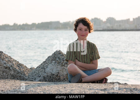 Porträt eines Jungen sitzen auf einem betonpfeiler auf dem Hintergrund des Meeres und die Strahlen der Sonne. Stockfoto