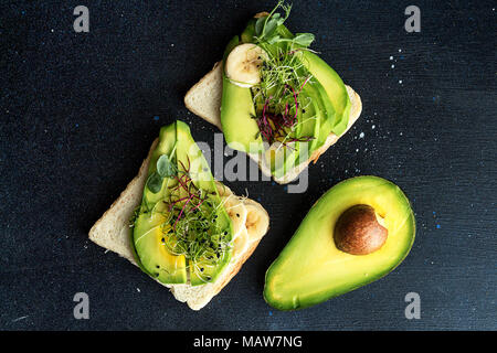 Gesundes Frühstück Toast mit Frischkäse, Scheiben Avocado, Banane und microgreen auf dunklem Hintergrund. Sauber Essen vegan Diät Konzept. Ansicht von oben. Stockfoto