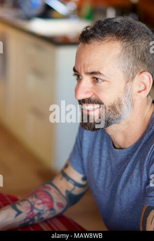 Von Mitte 30 Bart und tätowierte Mann lächelnd am Küchentisch mit einer roten Tischdecke und trägt ein blaues T-Shirt und ein großes Ansehen. Stockfoto