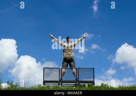Mann, der auf einer Bank, die Hände in Triumph, blauer Himmel mit weißen Wolken im Hintergrund Stockfoto