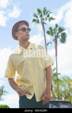 Ein junger kaukasischer Mann mit Hut, Sonnenbrille und gelben Hemd, posiert mit Palmen und blauem Himmel im Hintergrund. Eine männliche Urlaub lifestyle Konzept Stockfoto