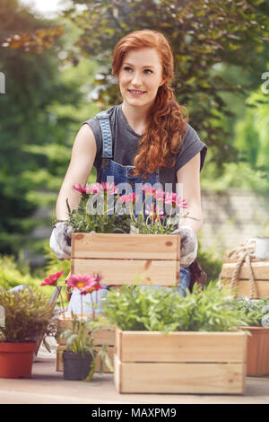 Lächelnde Mädchen in Arbeitshose, Verschieben eine Holzkiste mit Blumen und Pflanzen im Garten Stockfoto