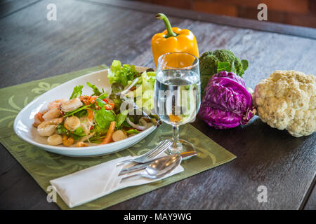 Schüssel Salat im Thai Stil mit Besteck, Glas Wasser einschließlich Brokkoli, Blumenkohl, Kohl und Paprika auf hölzernen Tisch im Restaurant Stockfoto