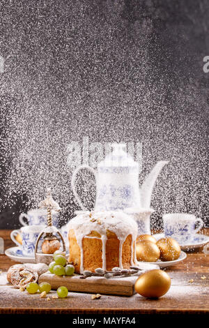 Schöne Karte für Ostern. Zucker Pulver wird auf die Ostern Kuchen gegossen. Leckereien für den Urlaub. Ostern Kuchen und goldene Eier mit einer Teekanne und Teetassen. Stockfoto