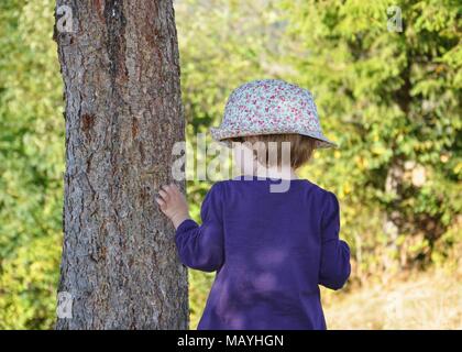 Zurück Seite Portrait von ein Kind, ein wenig blondes Mädchen mit Hut, berühren einen Baum im Wald Stockfoto