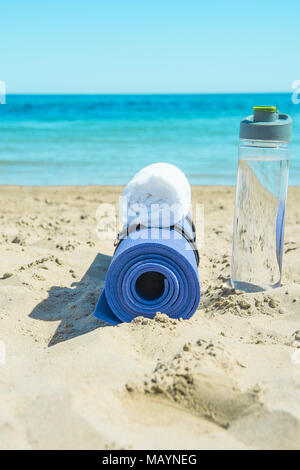 Yoga Matte rollte die Flasche mit Wasser Weiß Handtuch am Strand Sand und türkisfarbenem Meer blauer Himmel im Hintergrund. Sonnenlicht. Entspannung Sommer Meditation Fitness Stockfoto