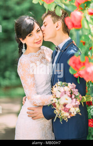 Brustbild des Bräutigams küssen die hübsche Braut in die Wange in die schöne rote Rosen Garten. Stockfoto