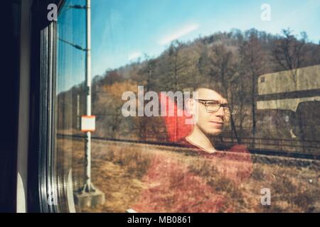 Transport auf der Schiene in sonniger Tag. Nachdenklicher junger Mann gesehen Obwohl Zug Fenster mit Reflexion auf Reisen. Stockfoto