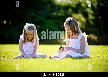 Zwei junge hübsche Caucansian Mädchen in weißen Röcken sitzen auf Gras spielen an einem Park mit Hintergrundbeleuchtung und verschwommen grünen Hintergrund Stockfoto